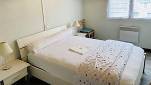 A neatly made bed with white linens and a star-patterned blanket, a bedside table with a lamp, a chair with a blue cushion, and a window with a lace curtain. - Small country house with ocean and shops on foot (045) (Biscarrosse)