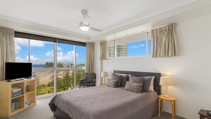Une chambre d’hôtel avec un grand lit, un téléviseur posé sur un meuble en bois, une bibliothèque, une table de chevet avec une lampe, et une vue sur la plage et la ville grâce à de grandes fenêtres.
