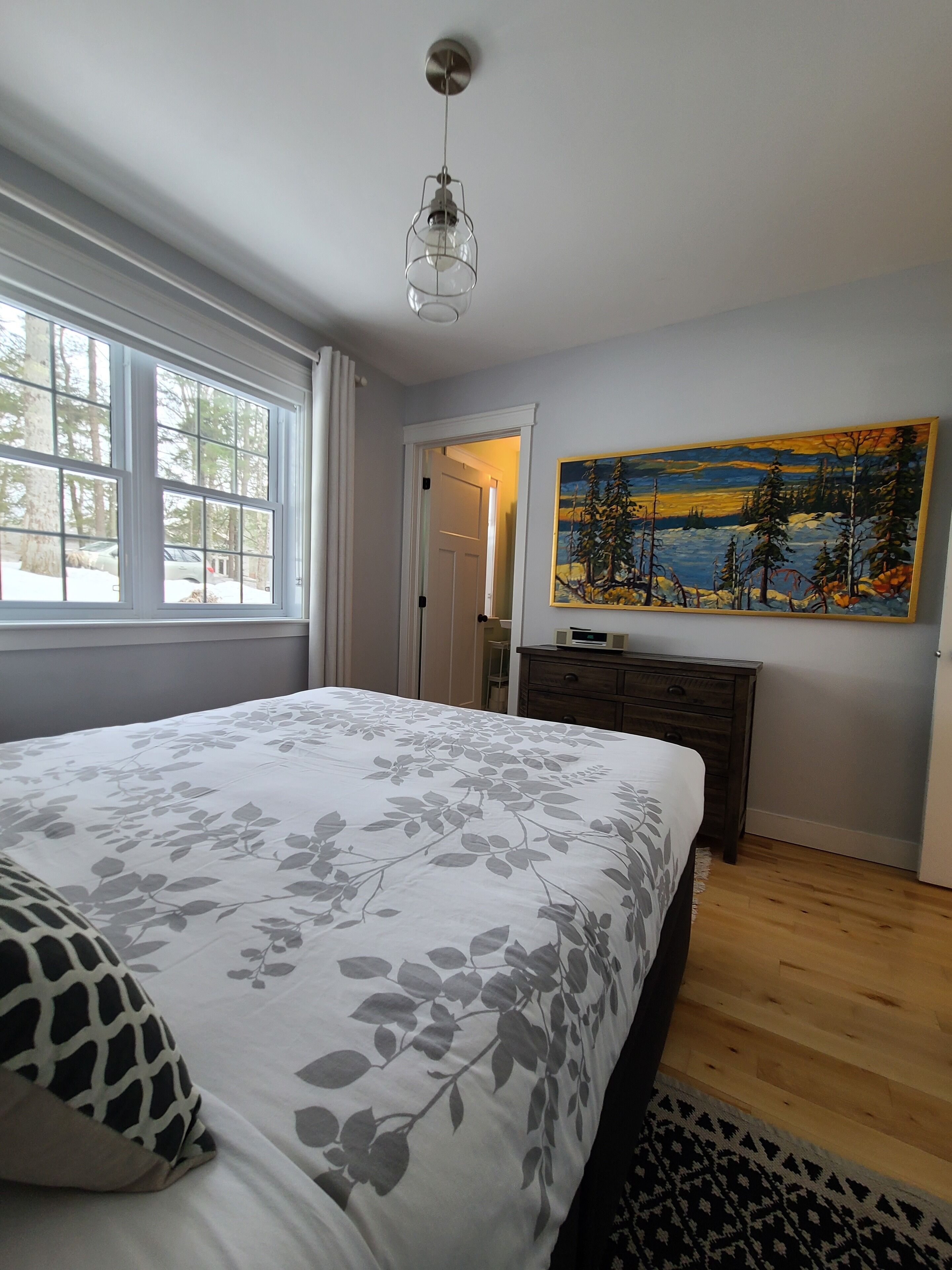 Ein Schlafzimmer mit einem großen Bett, einem Bild an der Wand und einem Fenster mit Blick auf Bäume.