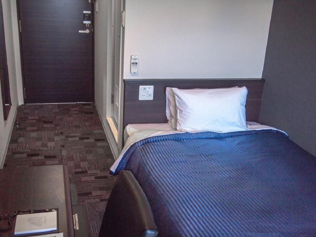 Una camera d'albergo con un letto, un comodino, una sedia, una porta e un pannello di controllo a parete.