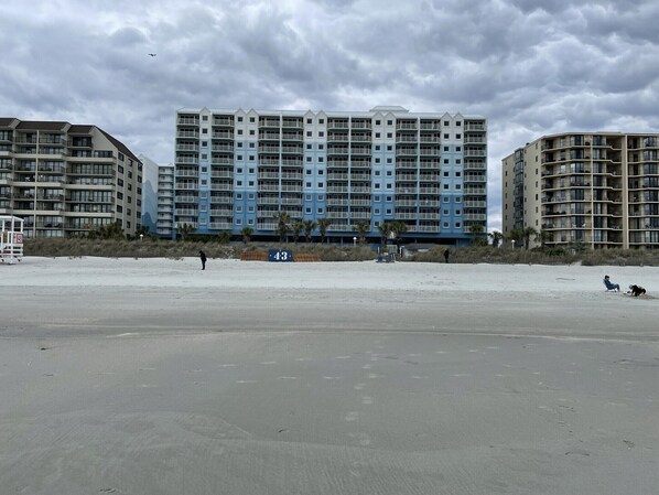A beachfront hotel with multiple floors, balconies, and a sandy beach in the foreground.