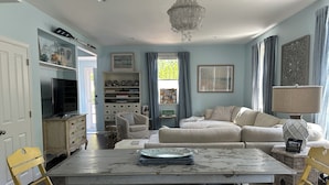 A living room with a sofa, a coffee table, and a chandelier. There is a television on a cabinet and a bookshelf with various items.