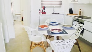 A compact kitchen with white cabinets, a dining table set for four, and a small window with blinds. - The Buddha House Torrox Costa (Torrox Costa)