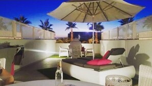 A patio area with a white sofa, a red cushion, and a table with a bottle. There is a large umbrella providing shade. - The Buddha House Torrox Costa (Torrox Costa)