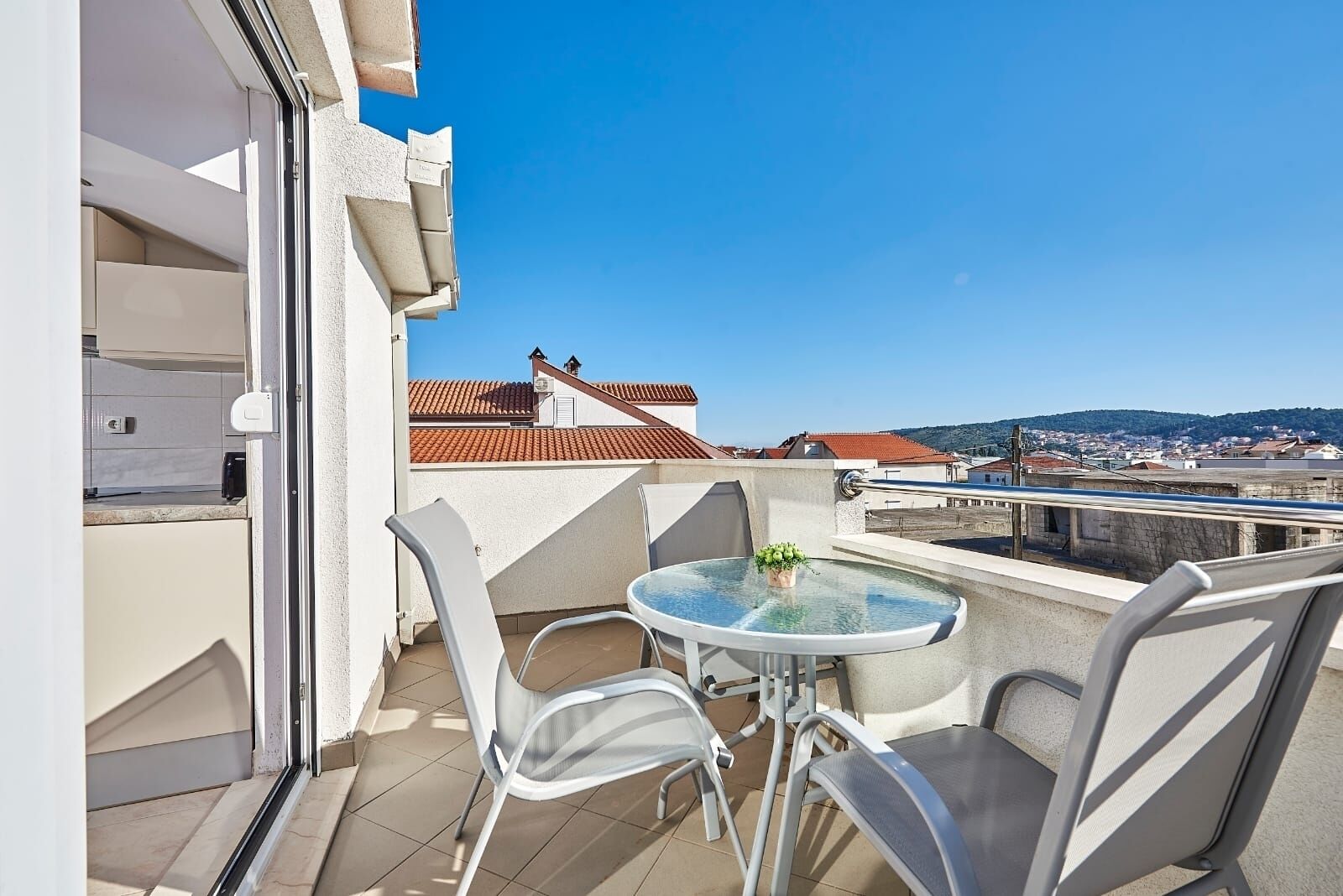 A balcony with a glass table and chairs, a small potted plant, and a view of buildings and hills in the distance.