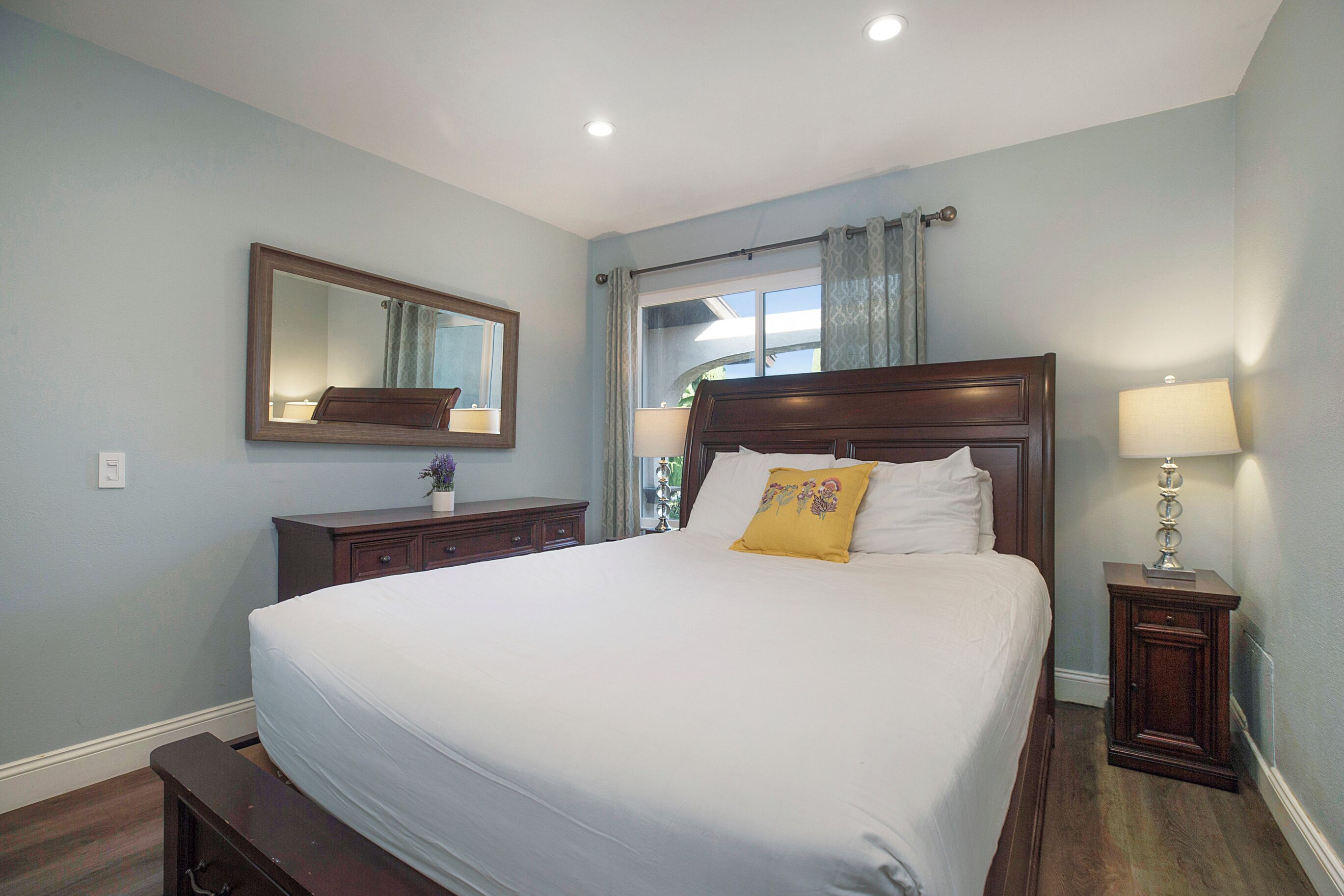 Una cama bien hecha con sábanas blancas y una única almohada amarilla, una mesita de noche de madera con una lámpara, un aparador con un espejo y una ventana con cortinas.