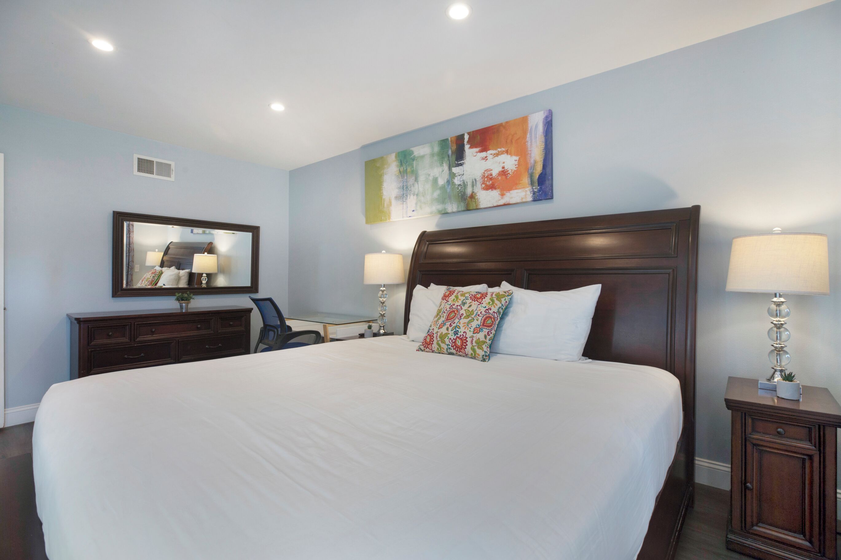 A hotel room with a large bed, two bedside lamps, a nightstand, a dresser, a desk, and a colorful abstract painting on the wall.