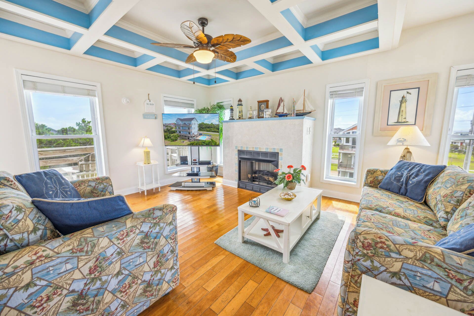 A spacious living room with a fireplace, two sofas with floral upholstery, a ceiling fan, and a large window offering a view of the outdoors.