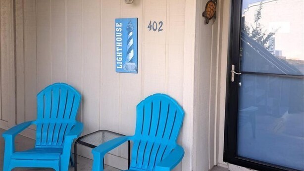 Un porche con dos sillas Adirondack azules, un felpudo, una puerta con un letrero de número y un accesorio de luz fijado a la pared.