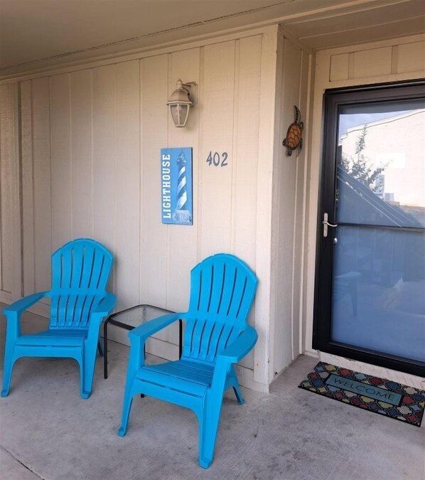 A porch with two blue Adirondack chairs, a welcome mat, a door with a number sign, and a wall-mounted light fixture.