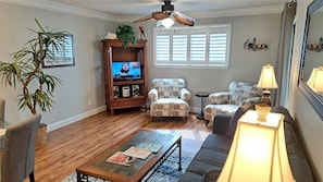 A living room with a wooden floor, two armchairs, a sofa, a coffee table, a TV, a ceiling fan, a window with blinds, and a lamp.