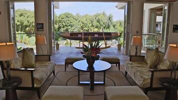 A spacious living area with a central round table, two sofas, and a coffee table. There are two lamps on either side of the table. The room has large windows offering a view of a garden and a pool.