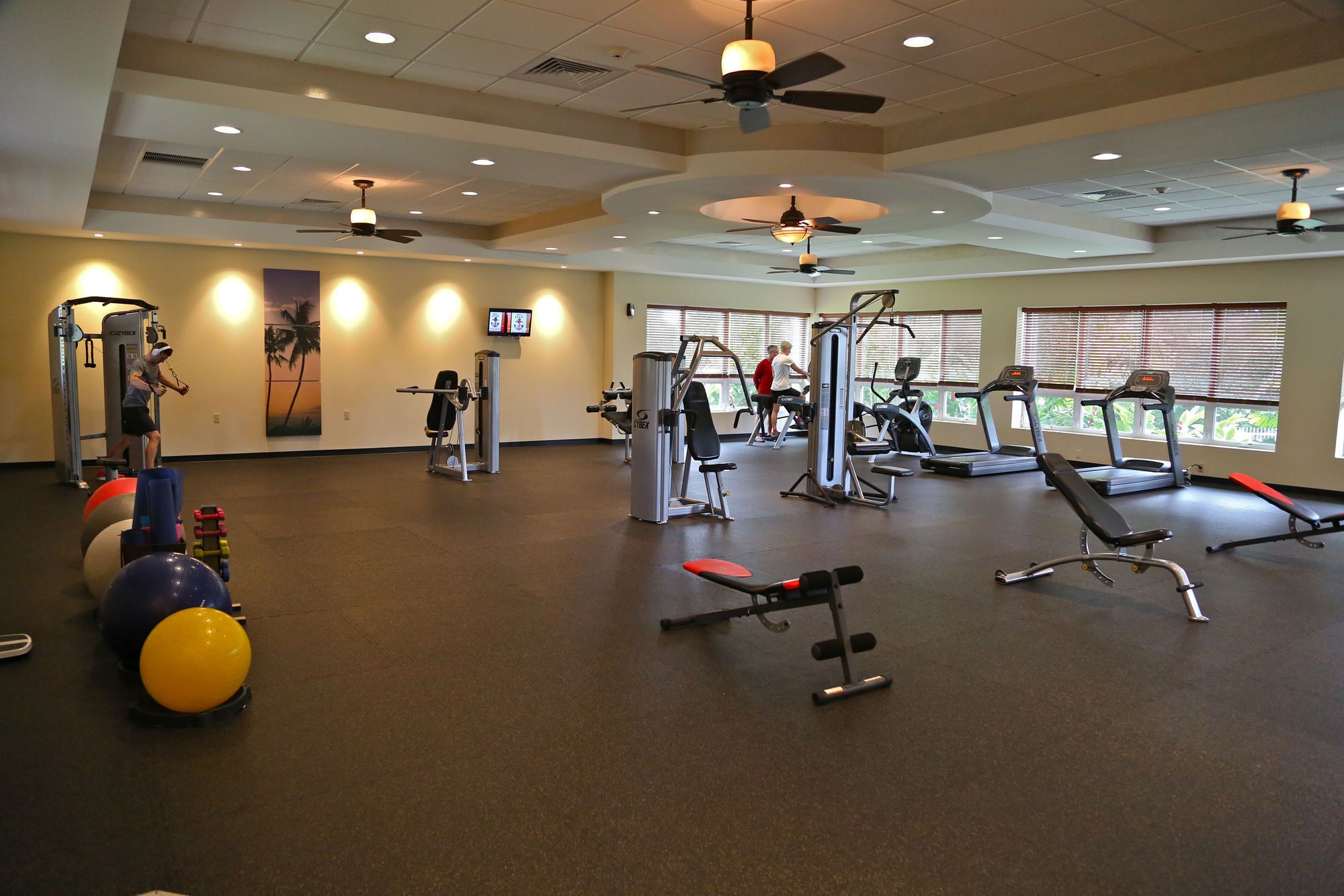 A gym with various exercise equipment, including treadmills and weight machines, and large windows providing natural light.