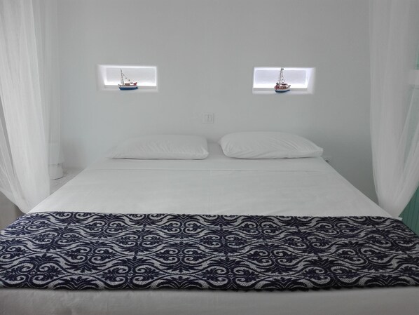 A bed with a white sheet, two pillows, and a patterned blanket. There are two small windows with curtains and a model ship in each.