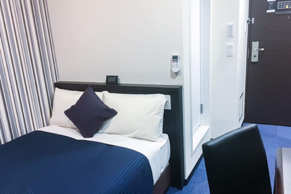 Een hotelkamer met een bed, nachtkastje, stoel, deur en een aan de muur bevestigd telefoon.