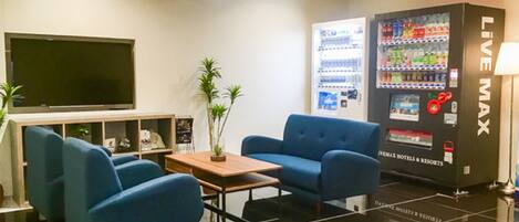 A modern hotel lobby with a flat-screen TV, a seating area with blue sofas, a wooden coffee table, and a vending machine labeled "LIVE MAX".