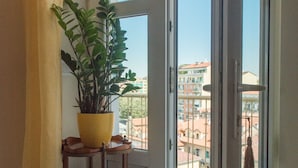A room with a large window, a potted plant on a wooden stand, and a curtain. - New picturesque and central penthouse, with a splendid view of the mountains (Torino)
