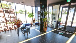 Uma área de recepção com piso de azulejos, balcão de atendimento, cadeiras, um vaso de plantas e uma porta de vidro que dá para o exterior.