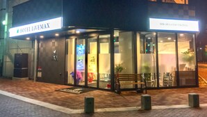 Entrada de um hotel com fachada de vidro, um banco na parte externa e uma placa com a inscrição 'HOTEL LIVEMAX'.