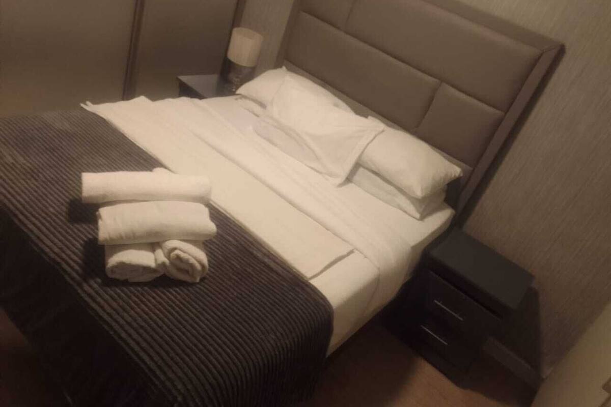 Una cama bien hecha con sábanas blancas, una colcha a rayas y toallas dobladas al lado. Hay una mesita de noche con una lámpara y un cajón.