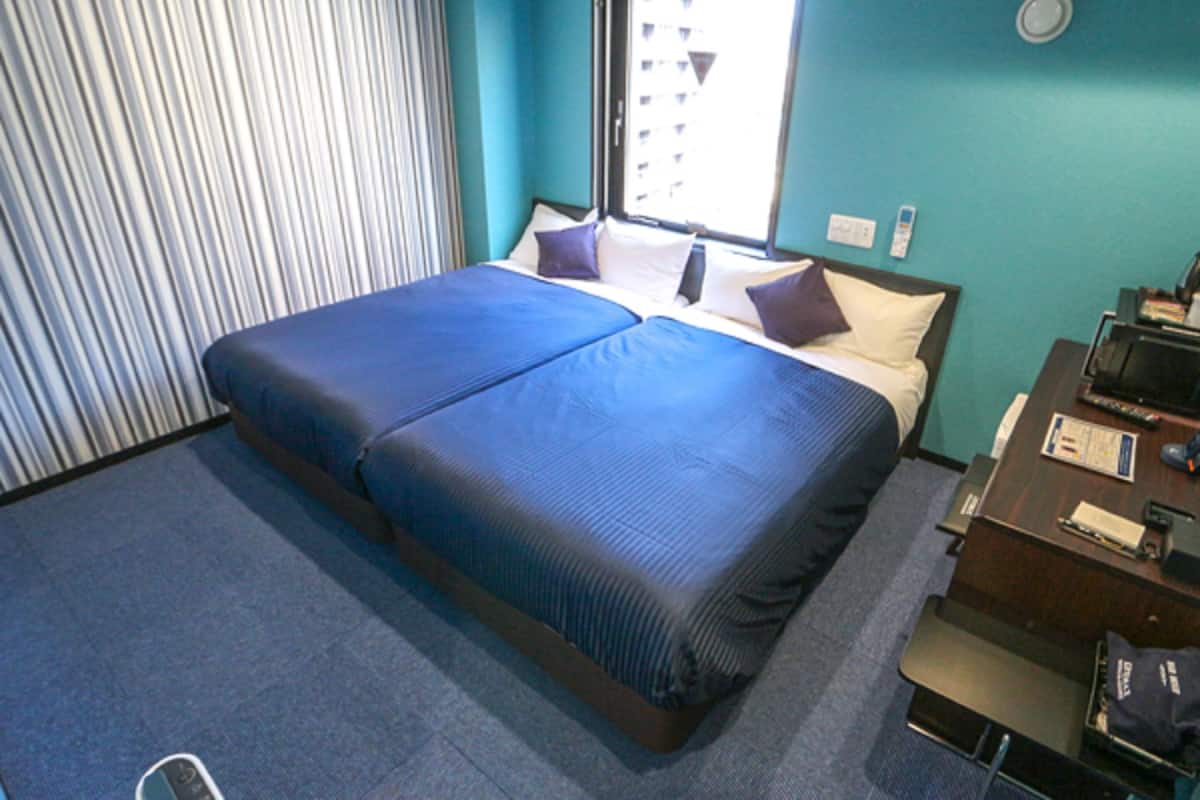 A hotel room with a large bed, a desk with a computer, and a window with blinds.