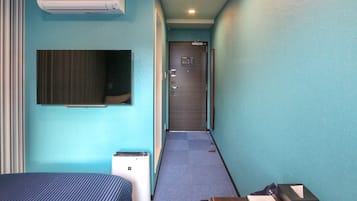 Habitación de hotel con cama, escritorio, silla, unidad de aire acondicionado, televisor y puerta que da a un pasillo.