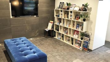 Una habitación con un banco acolchado azul, una estantería blanca con revistas y un televisor instalado en una pared gris.