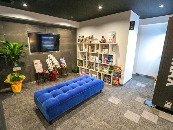 Un área de recepción con un sofá azul, una estantería con revistas, un televisor y una planta en maceta.