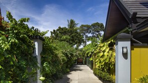 A narrow pathway flanked by lush greenery and a yellow building.