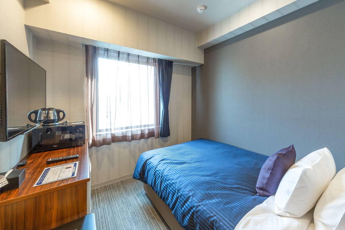 A hotel room with a bed, a desk with a computer, a window with curtains, and a mirror on the wall.
