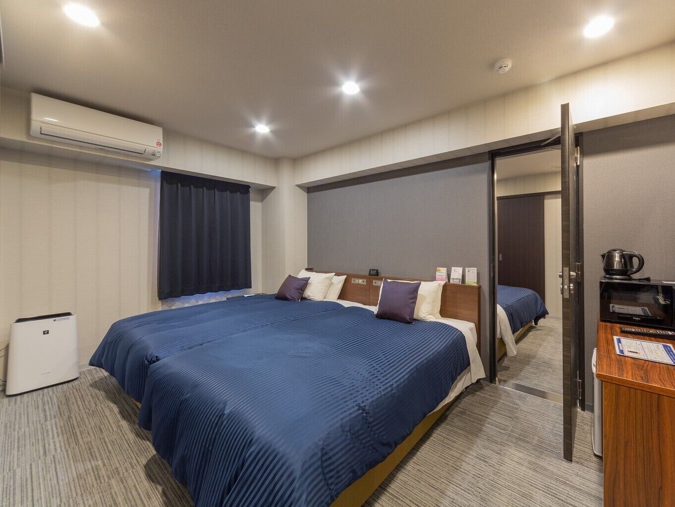 A hotel room with a large bed, a desk with a kettle, and an air conditioning unit.
