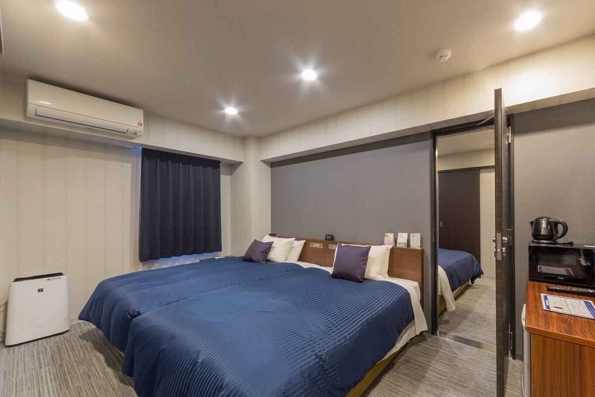 A hotel room with a large bed, a desk with a kettle, and an air conditioning unit.