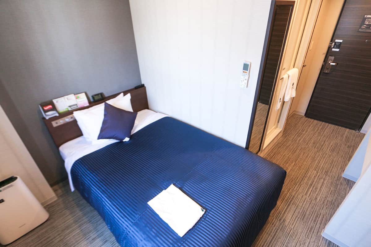 A hotel room with a bed, a nightstand, a control panel, and a door.