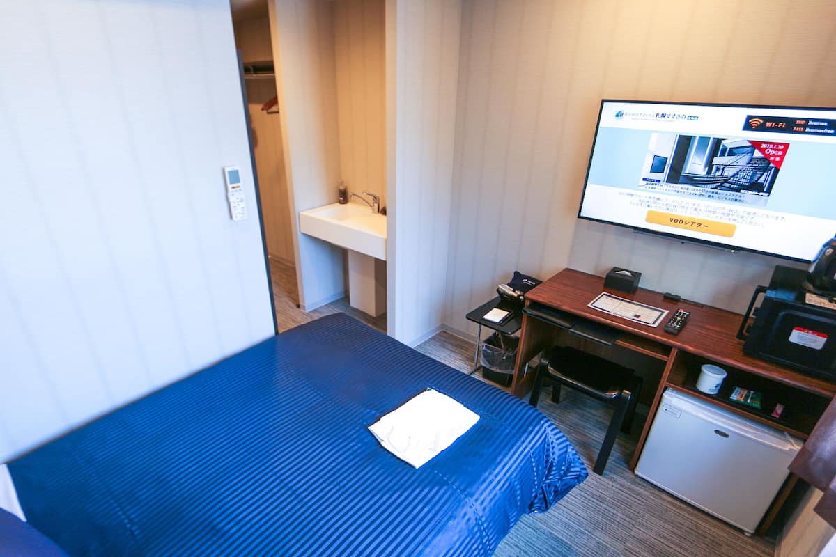 A hotel room with a single bed, a desk with a computer, a small fridge, and a TV displaying a webpage.