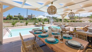 A poolside dining area with a wooden table set for six, surrounded by wicker chairs, under a canopy with hanging rattan lamps. - Anesis Villa, with Private Pool & BBQ, Walking distance to the beach & amenities (Rethymno)