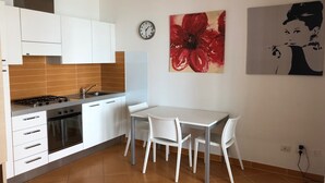 Een moderne keuken met witte kastjes, een gasfornuis en een gootsteen. Er is een eettafel met vier stoelen en een klok aan de muur.