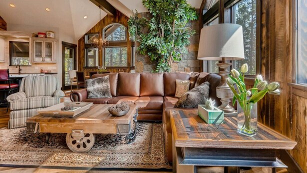 A spacious living room with a large brown leather sofa, a coffee table, and a rug. There is a dining area with a table and chairs, and a kitchen in the background.