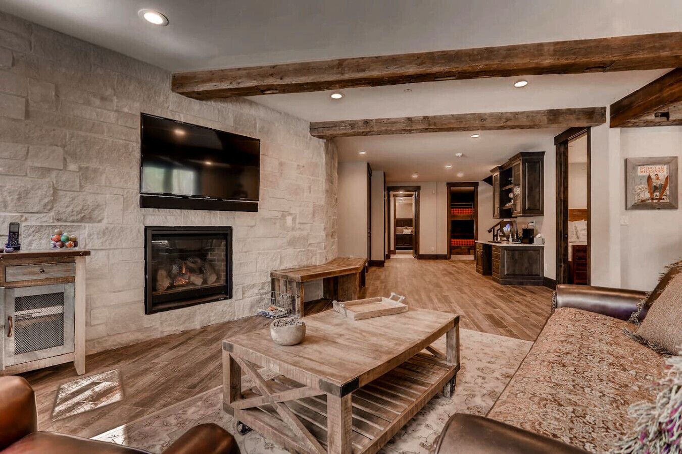 A spacious living room with a stone fireplace, wooden beams, a TV mounted on the wall, and a cozy seating area.