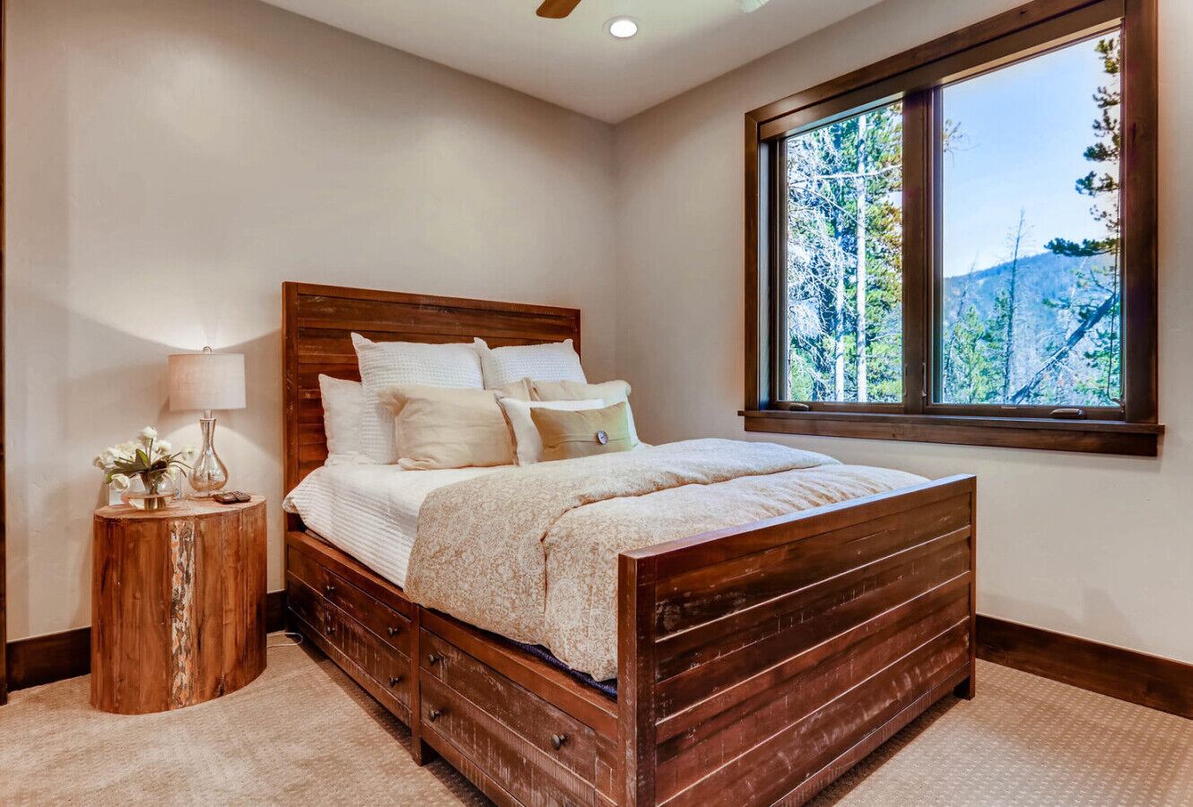 Quarto com cama de madeira, mesa de cabeceira, luminária e uma grande janela com vista para árvores e colinas.