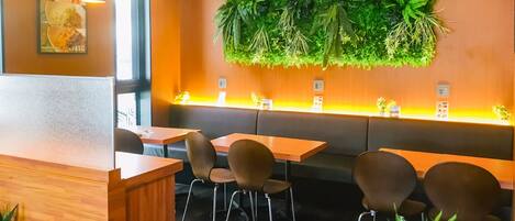 A restaurant interior with wooden tables and chairs, a green plant wall, and a menu poster on the wall.