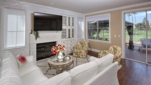 A spacious living room with a white sofa, a fireplace, a television, and a view of a golf course through large windows. - Stunning golf course view vacation home (Tustin)