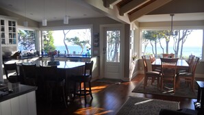 A spacious kitchen with a large island, wooden dining table, and chairs. There are multiple windows offering views of trees and the ocean. -  Secluded Sandy Beach All To Yourself! Panoramic Ocean and Mountain views. (Comox)