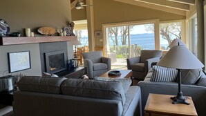 A living room with a fireplace, grey sofas, a coffee table, and a view of the sea through large windows. -  Secluded Sandy Beach All To Yourself! Panoramic Ocean and Mountain views. (Comox)