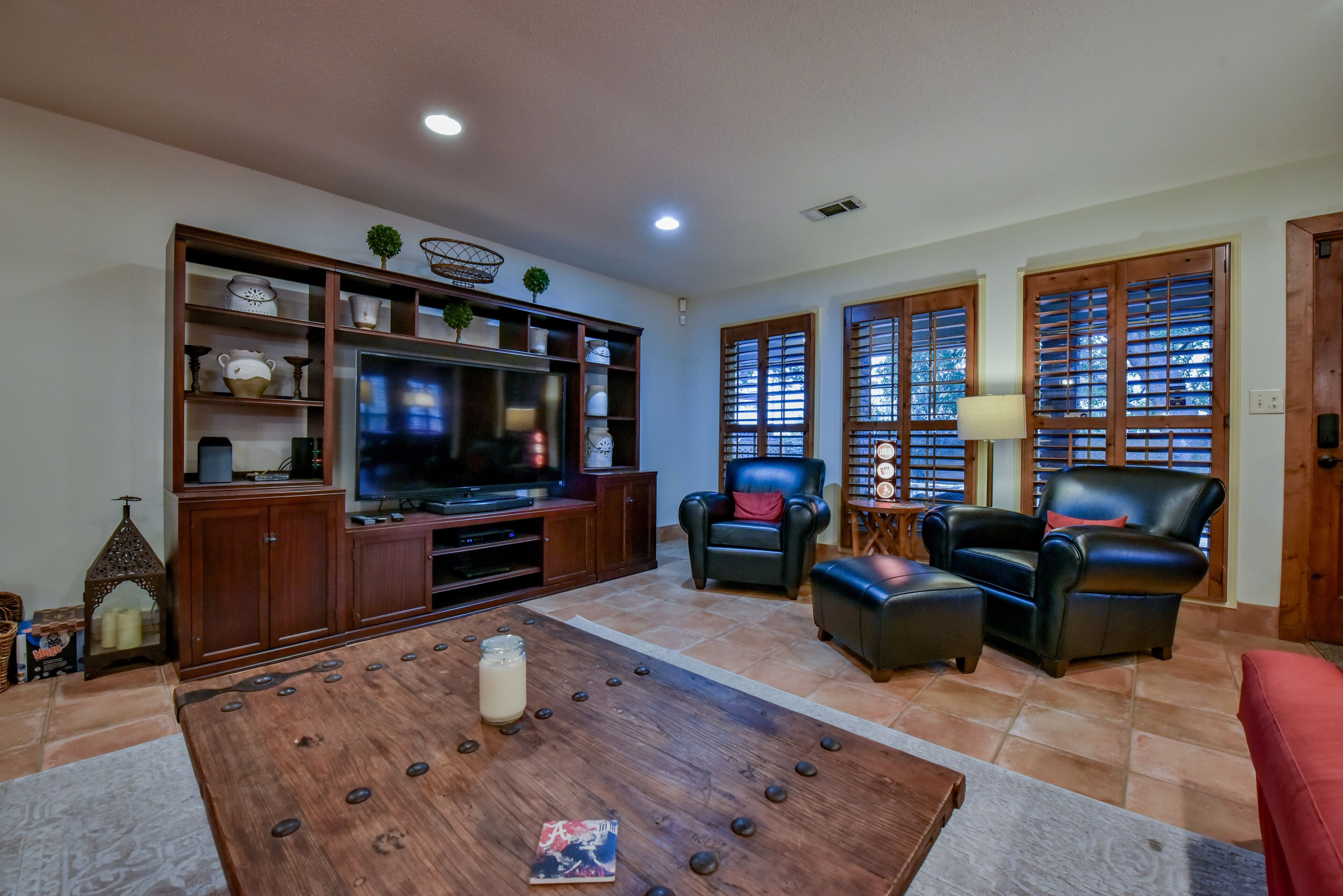 A living room with a wooden entertainment center, a flat-screen TV, leather chairs, a coffee table, and a window with wooden shutters.
