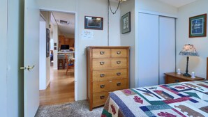 Une chambre avec une commode en bois, un lit recouvert d’une courtepointe, un bureau avec une lampe et une porte donnant sur une autre pièce.