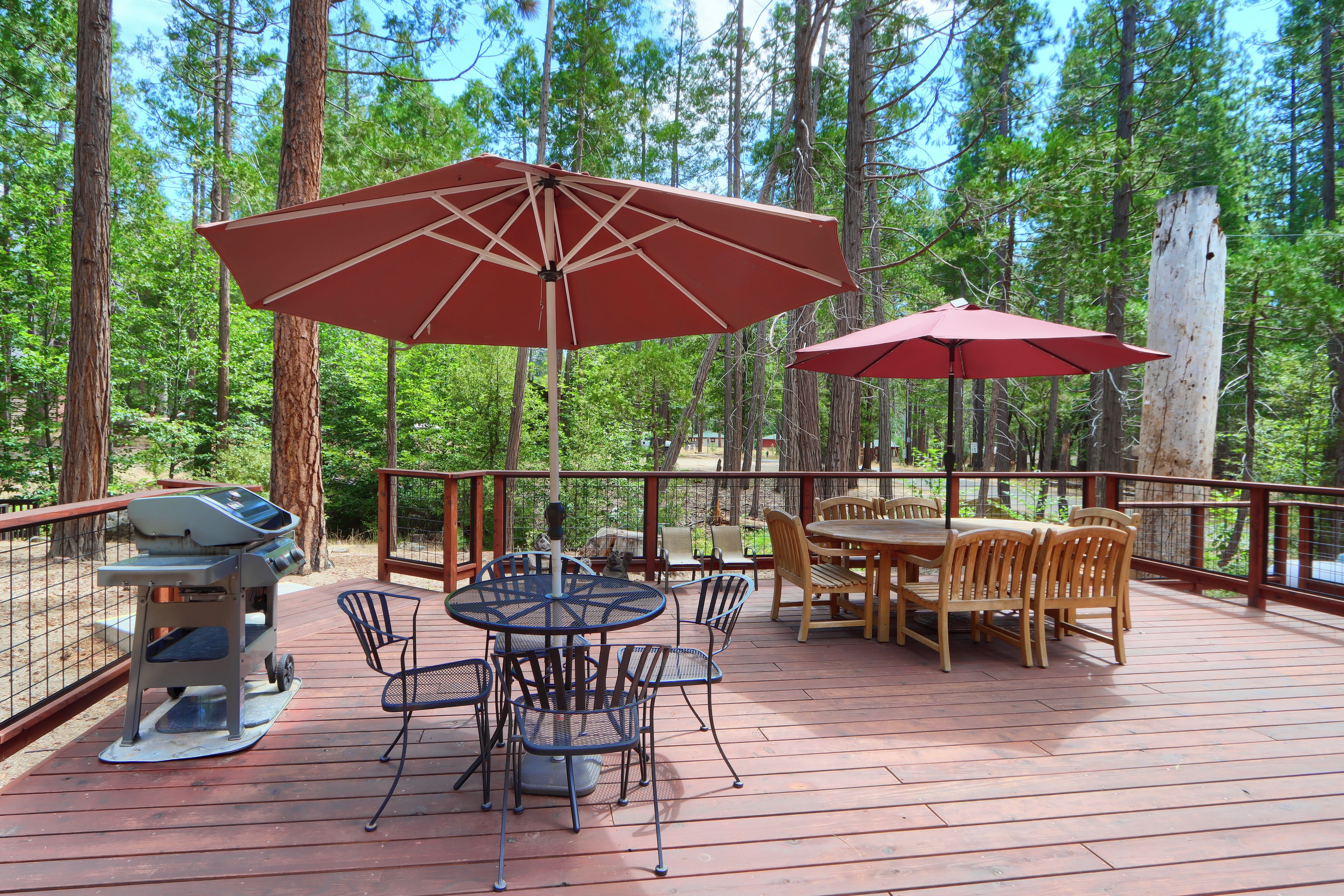 Une terrasse en bois avec un barbecue, une table ronde et des chaises, entourée de parasols rouges et offrant une vue sur de grands arbres.