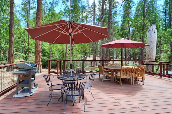 Eine Holzterrasse mit Grill, rundem Tisch und Stühlen, umgeben von roten Sonnenschirmen und Blick auf hohe Bäume.
