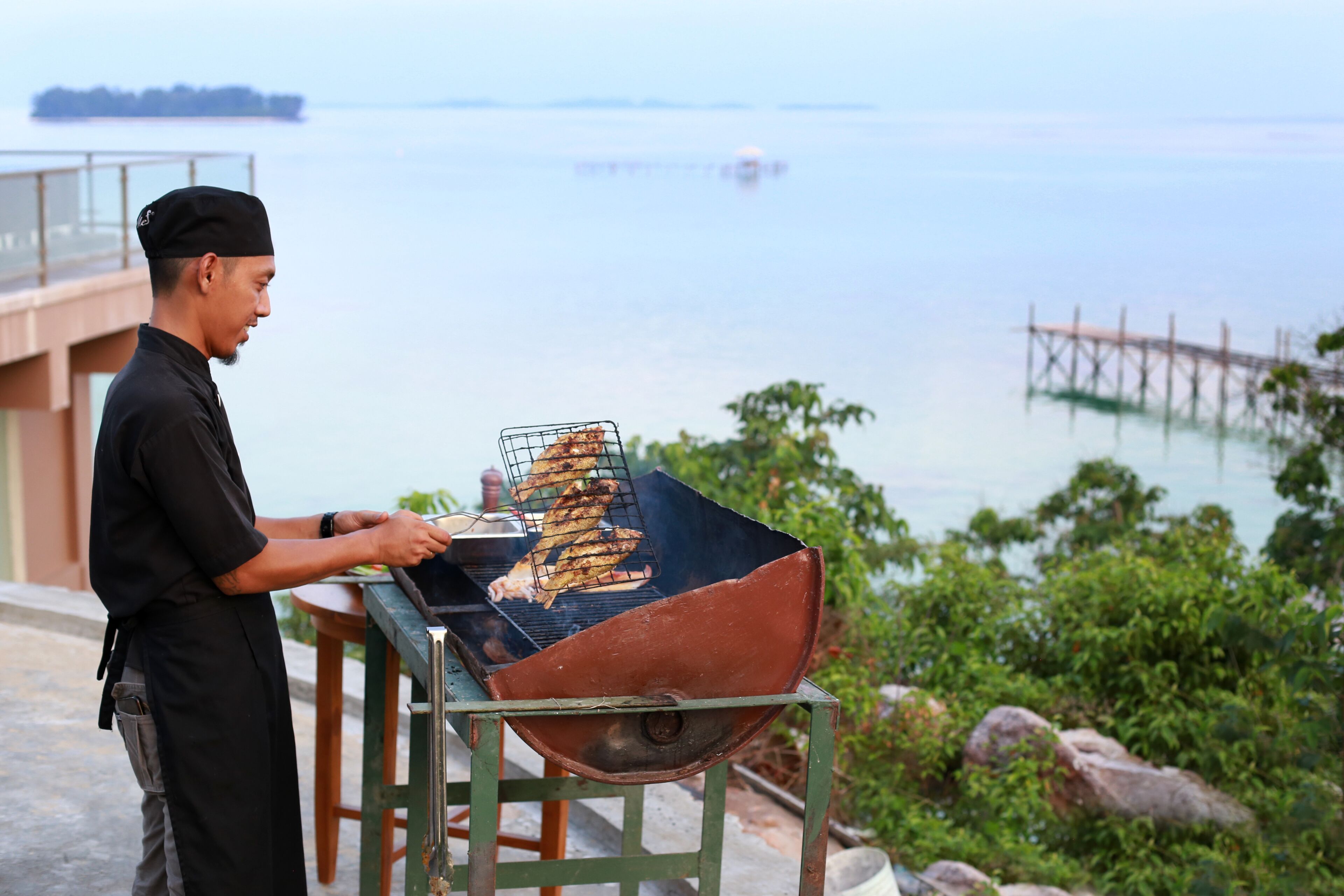 A person grilling outdoors with a view of the sea and a wooden pier in the background.