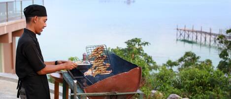 A person grilling outdoors with a view of the sea and a wooden pier in the background.