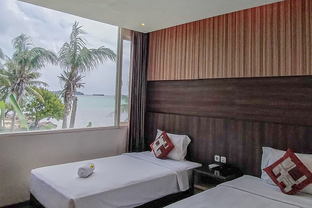 A hotel room with two beds, a window with a view of palm trees and the sea, and a wooden headboard.